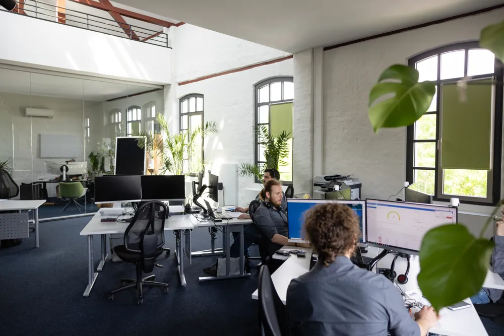 Großraumbüro mit Rundbogenfenstern, Pflanzen und Arbeitsplätzen des Technik-Teams